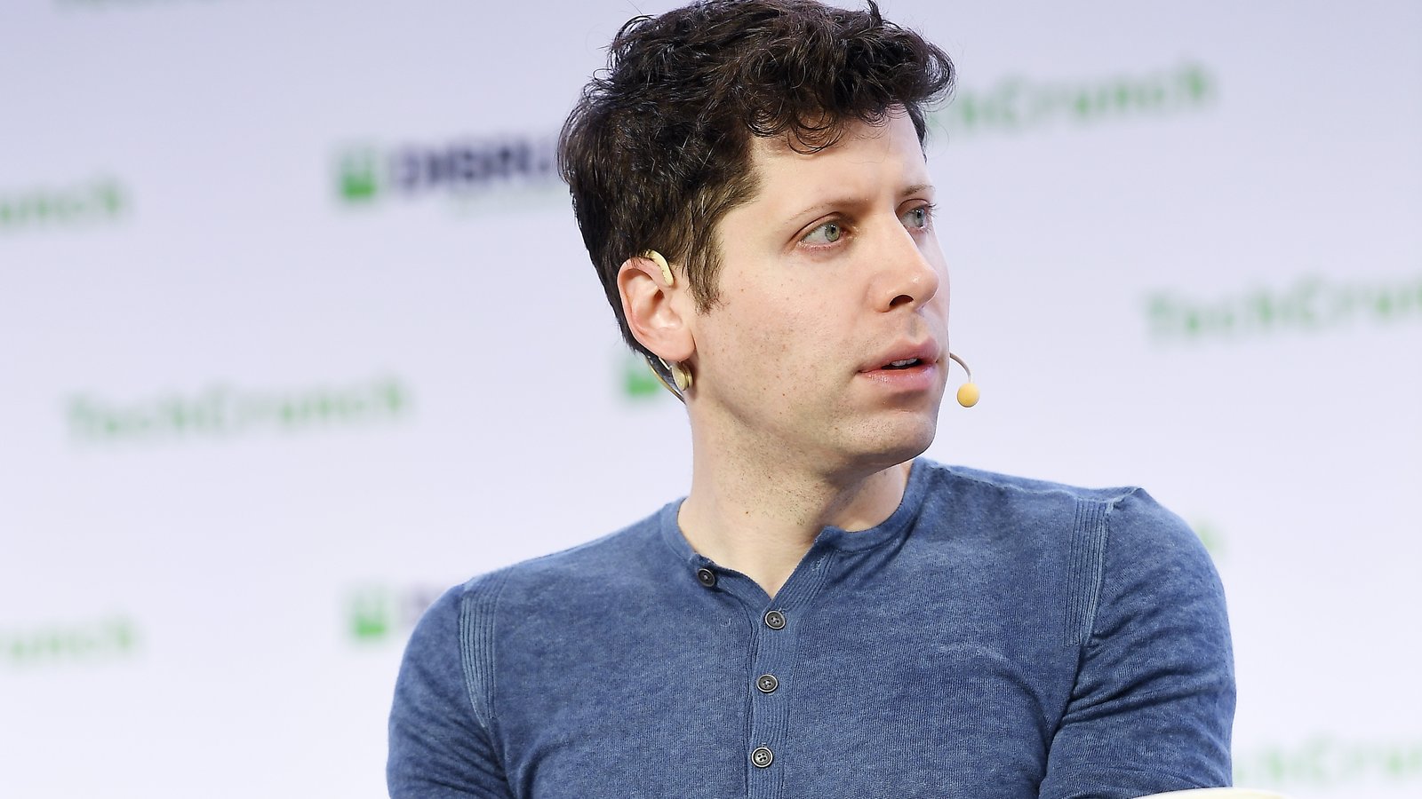 Sam Altman mặc áo xanh ngồi trên ghế sân khấu TechCrunch Disrupt, đeo micro cài tai, nhìn sang bên phải